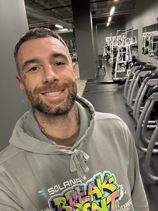 A man smiles for a selfie in a gym, wearing a gray hoodie that reads "SOLANA BREAKPOINT," tying into his post about prioritizing gym time ("Health is wealth!") while also working on the upcoming Solana Breakpoint conference.