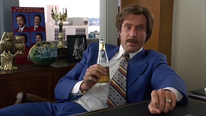 Will Ferrell as Ron Burgundy in a blue suit holding a beer bottle, sitting in an office with a 'RON BURGUNDY: #1 IN SAN DIEGO' poster behind him.