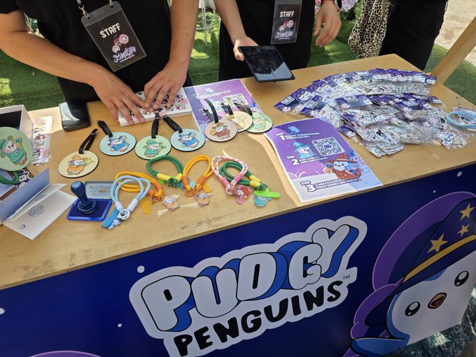 A vendor table at a Pudgy Penguins promotional event, displaying branded merchandise including bags, keychains, badges, and cards, with staff wearing 'STAFF' lanyards.