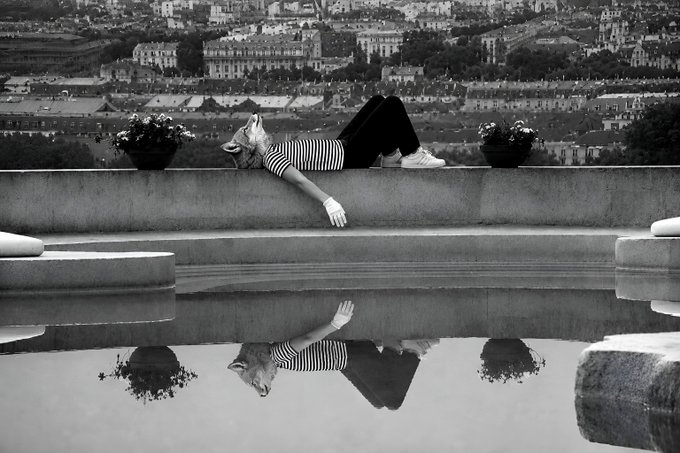 A black-and-white photograph of a woman in a striped shirt reclining on a stone ledge overlooking a cityscape, with her reflection mirrored in a pool of water below.