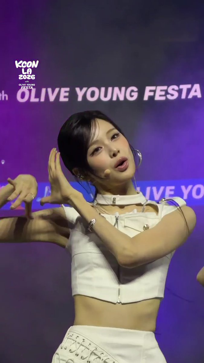 A performer from the K-pop group izna poses dynamically on stage with hands raised, wearing a white cropped top and skirt at the KCON LA 2026 Countdown Stage, as indicated by the "KCON LA 2026 with OLIVE YOUNG FESTA" branding visible in the background.