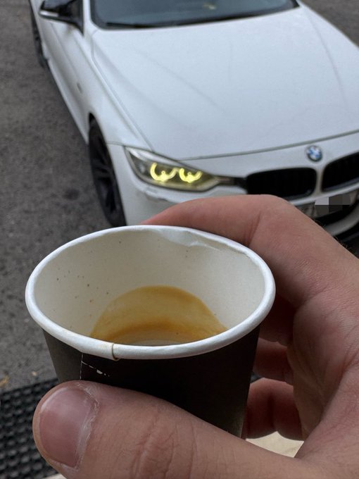 A hand holding a nearly empty paper coffee cup in the foreground, with a white BMW car with its headlights on parked in the background.