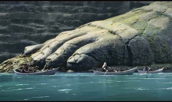 An animated scene depicting three small rowboats carrying people across turquoise water near a massive rocky cliff or stone giant's hand.