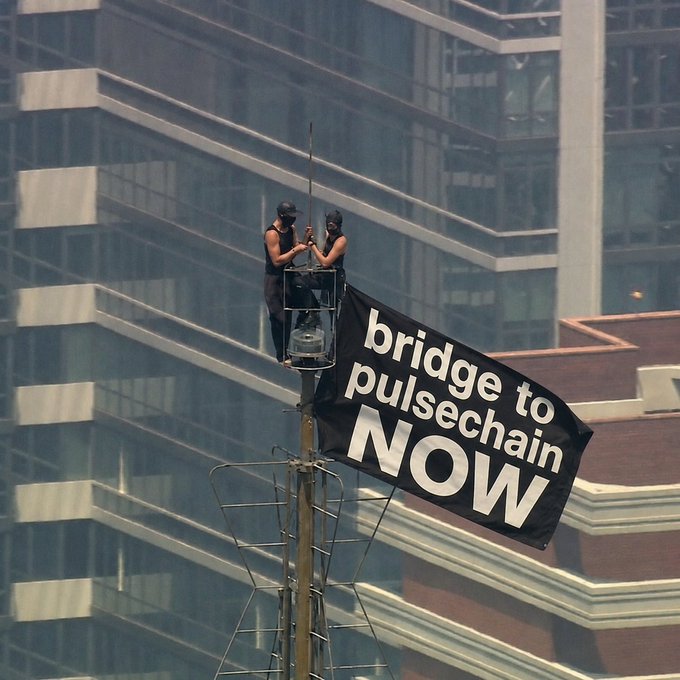 Two masked individuals stand in a small platform attached to a high structure, with a black flag reading 'bridge to pulsechain NOW' attached below them, set against a city skyline.
