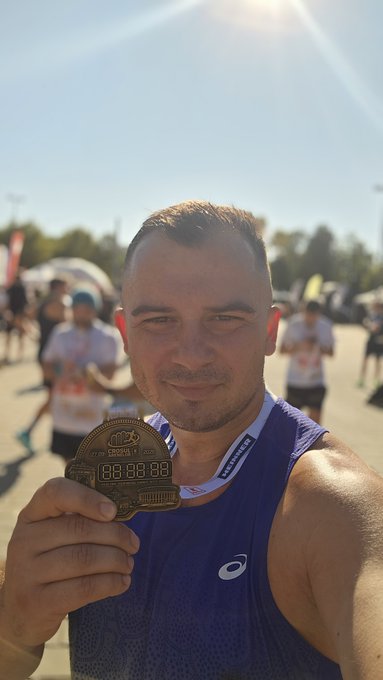 A man in a blue ASICS athletic tank top holds up a medal at what appears to be a running event, with a bright sunny sky and other participants in the background.