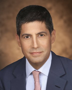 A professional headshot of Kevin Warsh, a former Federal Reserve governor, wearing a navy suit, light blue shirt, and pink tie. The image accompanies his quoted statement comparing Bitcoin to gold for younger investors.