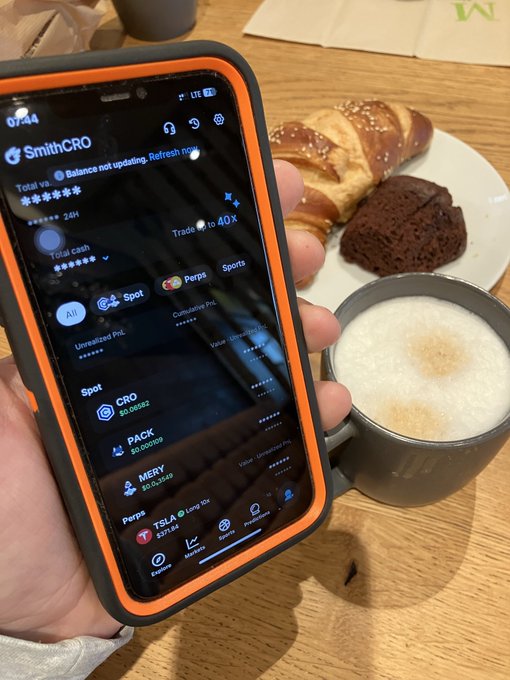 A hand holds a smartphone displaying the SmithCRO crypto trading app showing balances for CRO, PACK, MEMH, and FSLA, with a plate of pastries and a cup of latte visible on the table nearby.