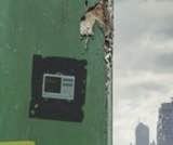 A black electronic device or breaker box mounted on a green metal panel, with a city skyline and cloudy sky visible in the background.