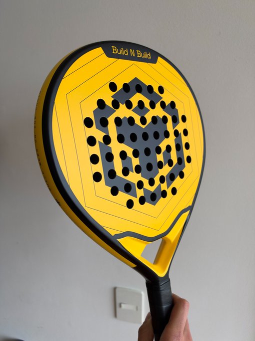 A close-up of a yellow and black padel/pickleball racket branded 'Build N Build' being held up against a white wall.