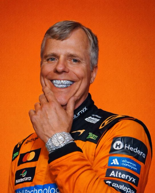 A smiling man wearing an orange McLaren racing suit with sponsor logos including Hedera, Alteryx, and Mastercard, posing with his hand on his chin against an orange background.