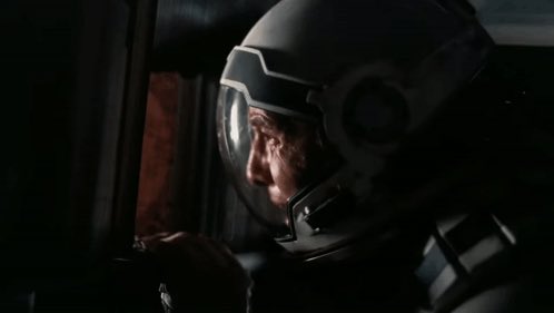 A cinematic close-up of an astronaut wearing a white space helmet with a reflective visor, sitting inside a dark spacecraft interior.