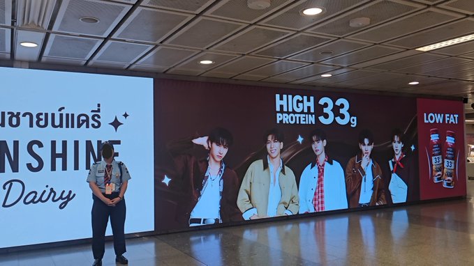A security guard stands in front of a large advertisement featuring five young men promoting a high-protein, low-fat dairy product by 'Sunshine Dairy' with '33g' prominently displayed.