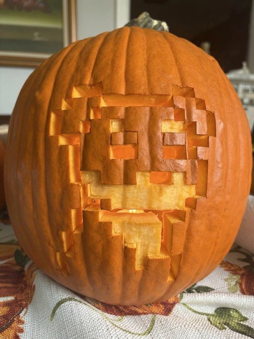 A carved orange pumpkin with an illuminated pixel-art skull design glows from within on a patterned tablecloth.