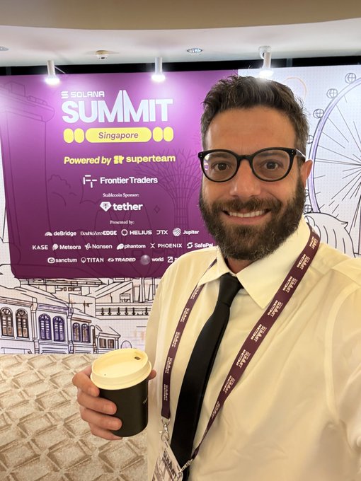 A man wearing glasses, a white shirt, black tie, and a lanyard holds a coffee cup while smiling at the camera in front of a 'SOLANA SUMMIT Singapore' banner powered by Superteam.