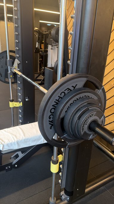 A loaded barbell with black "Technogym" weight plates sits on a Smith machine rack in a gym, ready for a heavy lifting session—perfect for those planning to "pump it."
