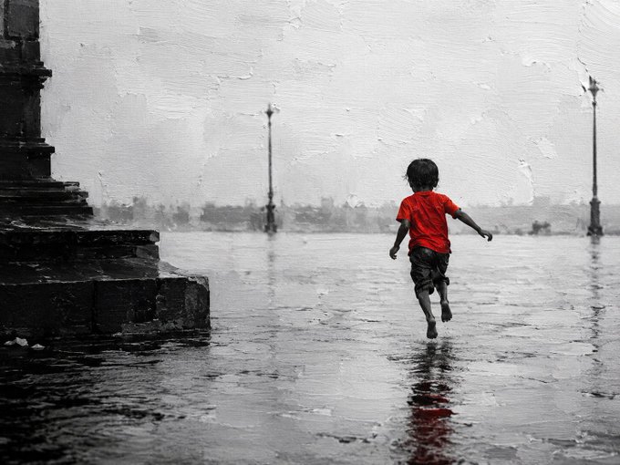 A child in a red shirt and dark shorts runs barefoot through a rain-soaked plaza, with a partially visible black-and-white painted structure on the left and lampposts in the distance.