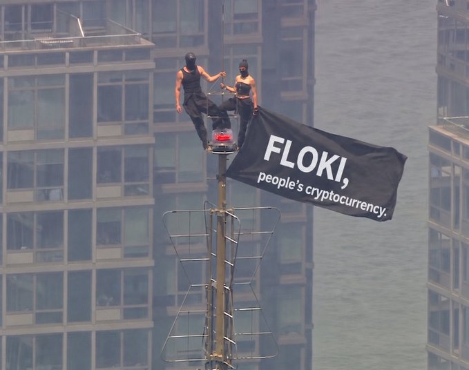 Two masked figures in black sit atop a tall building mast against a city skyline, holding a large black banner reading 'FLOKI, people's cryptocurrency.'