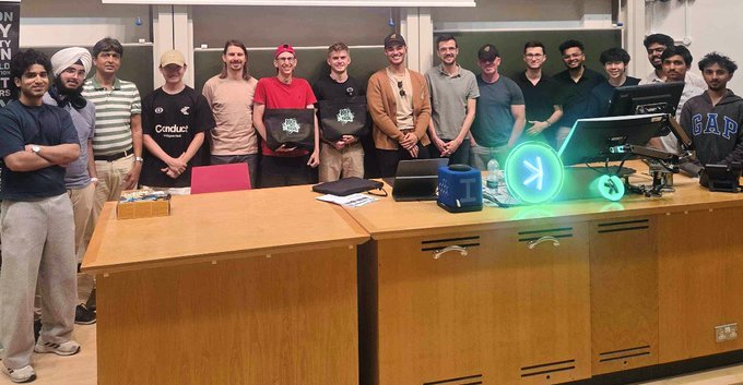 A group photo of approximately 15 people posing together in what appears to be an office or conference room setting, with a glowing Kaspa (K) logo displayed on a table in front of them.