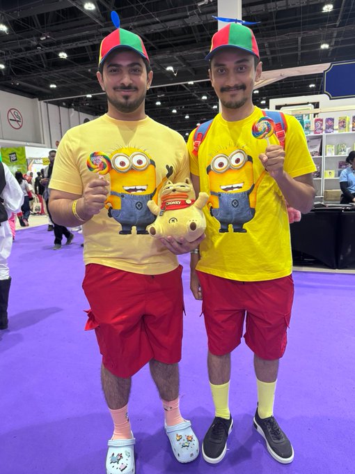 Two men dressed in matching Minion-themed costumes, complete with yellow shirts, red shorts, and propeller hats, pose with lollipops and a plush toy at what appears to be a convention.
