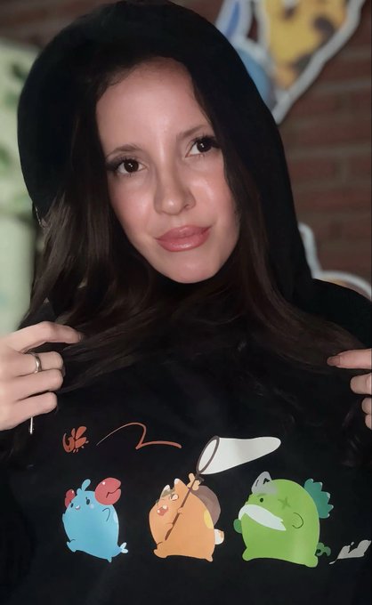 The image features a woman wearing a black hooded sweatshirt with colorful cartoon characters, including a frog, a bird, and a fish. The woman appears to be posing for a photo or participating in an event related to the Axie universe. The post also mentions that there is a new part of the Axie universe about to wake Terrariums at the AxieInfinity network.