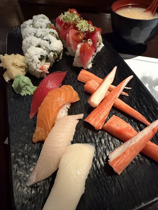 A black plate containing various types of sushi and nigiri, including salmon and crab sticks, accompanied by miso soup in a bowl.