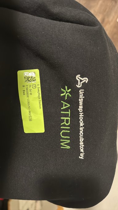 A close-up of a black garment with the text 'UniswapHook Incubator by ATRIUM' embroidered on it, along with a green tag containing a QR code and sizing details.