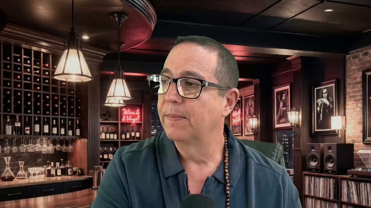 The image features a man wearing glasses, sitting at a bar with several bottles of wine behind him. He appears to be engaged in conversation or observing something off-camera. The background includes a dining table surrounded by chairs, suggesting that this scene takes place in a restaurant or similar establishment. The post is titled "Satellites To Blockchain To XRP youtu.be/_6PxXzuGuI4".