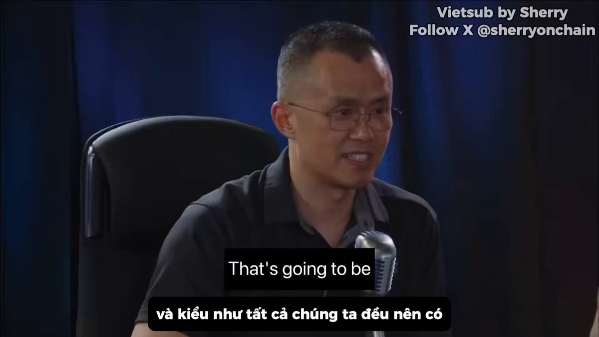 The image shows a man sitting at a desk with a microphone in front of him. The man appears to be speaking into the microphone, possibly giving a speech or presentation. The background features a blue curtain that partially covers the scene. The caption below the image reads "That's going to be CZ tiết lộ @binance đân khác hàng, quản lý rủi ro và vận hành."