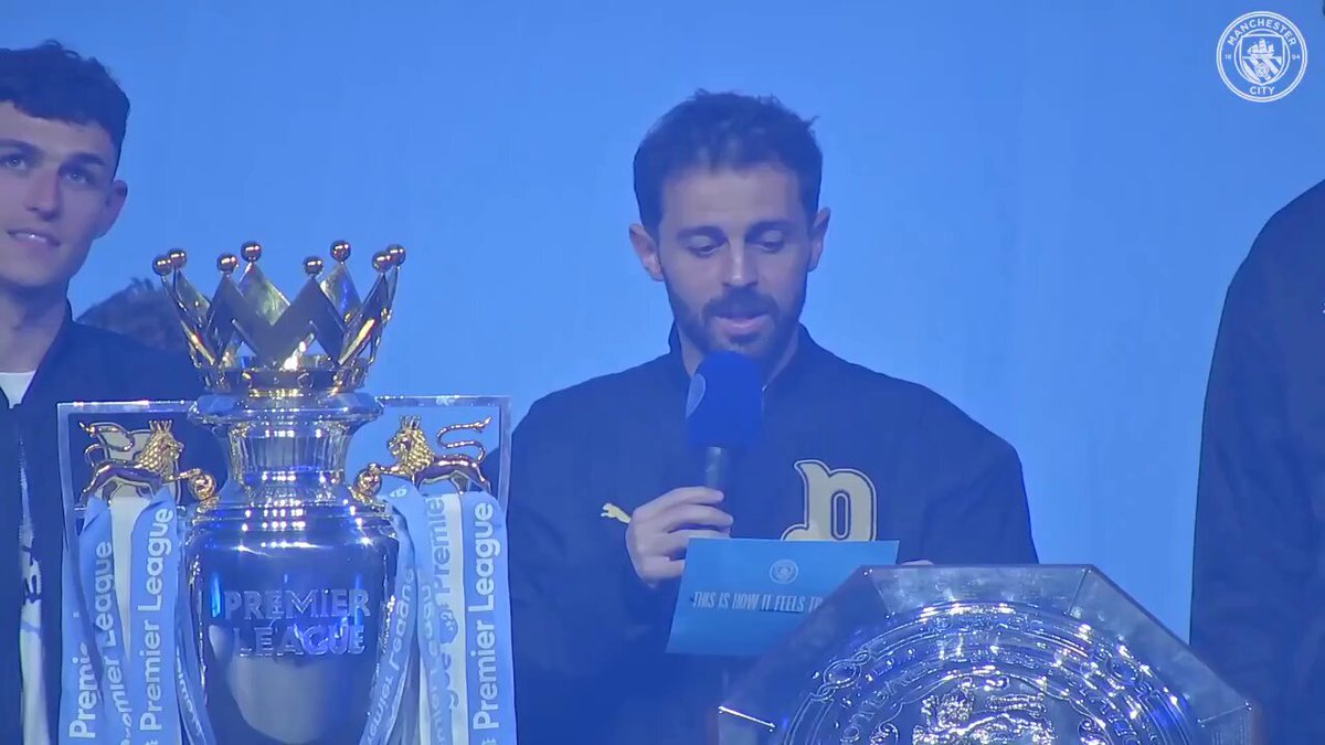 The image shows a man standing at a podium holding a trophy while giving a speech or presentation. The trophy has a crown-like design and is placed behind him. There are other people present, including one person sitting next to the podium and another person on the right side of the frame. A microphone can be seen in front of the speaker, indicating that he is addressing an audience during this event.