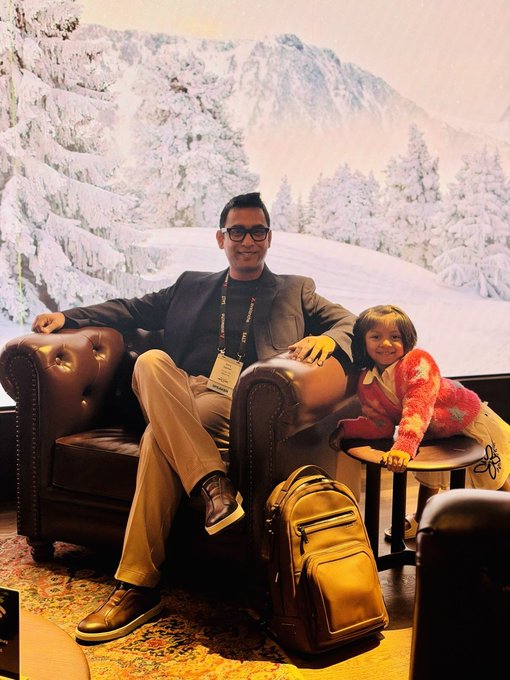 A man wearing glasses and a lanyard sits in a brown leather armchair next to a smiling young girl, with a tan backpack on the floor in front of them and a snowy mountain mural in the background.