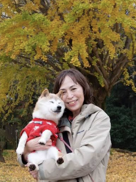 A woman with shoulder-length brown hair, wearing a beige jacket, holds a small Shiba Inu dog dressed in a red bandana amid autumn foliage with golden yellow leaves. The dog appears to be Kabosu, the famous Shiba Inu behind the Doge meme, connecting the image to Atsuko Sato's public statement distancing herself from the $CATE memecoin.