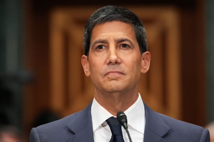 The image shows a man wearing a suit, tie, and speaking into a microphone at an event or gathering. He appears to be addressing an audience, possibly delivering a speech or presentation. The focus of the image is on the man's attire and his position in front of a crowd, rather than the content of what he is saying.
