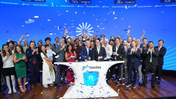 A group photo of people celebrating at what appears to be the Nasdaq stock market, with confetti falling and a screen displaying the Nasdaq logo and ticker symbols.