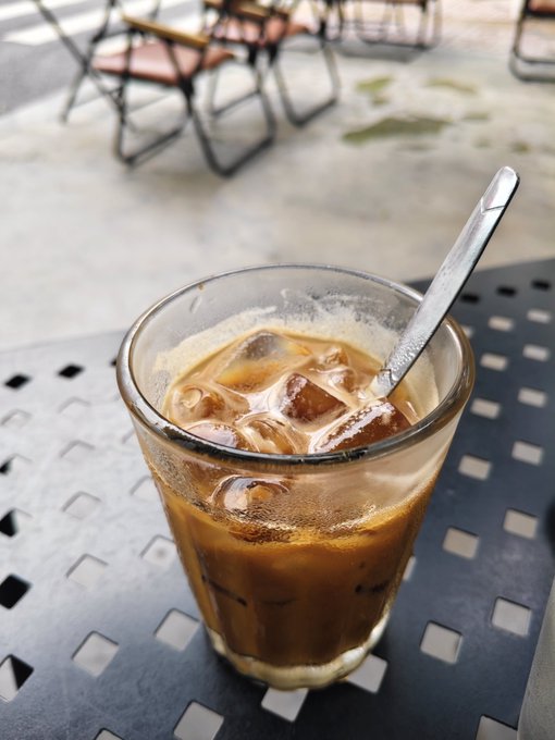 A glass of iced coffee with a metal spoon sits on a black metal patio table, with chairs visible in the blurred background.