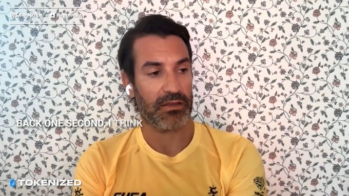 A man with a beard wearing a yellow shirt and earbuds sits in front of a floral wallpaper, with on-screen text reading 'BACK ONE SECOND, I THINK', a 'TOKENIZED' logo in the corner, and a stock ticker showing 'VISA Script', 'Alligent', and 'F5'.