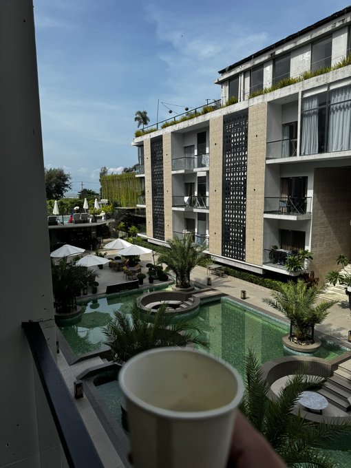 A view from a balcony overlooks a resort courtyard with a swimming pool, palm trees, white umbrellas, and a multi-story building, with a hand holding a cup in the foreground.