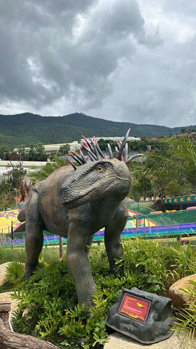 A large dinosaur statue with pink and blue spikes along its back stands in a grassy area with a small red informational sign, set against a backdrop of green hills and cloudy sky.