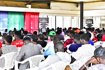 The image shows a large group of people sitting in chairs at an event with a banner that says "OKX" hanging above them, possibly indicating the name or theme of the gathering. The attendees are engaged and focused on the speaker who is standing behind them. The atmosphere appears lively and dynamic as everyone seems to be enjoying themselves and participating actively in the event.