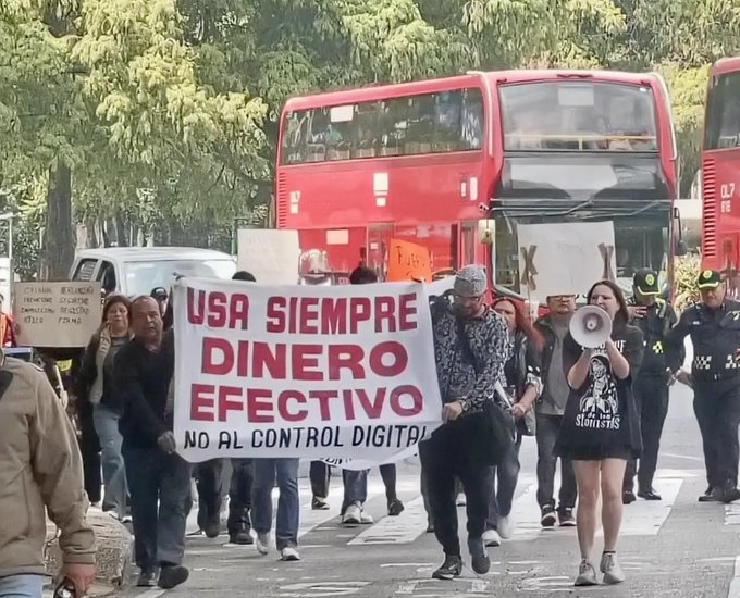 A protest scene where people march carrying a large banner reading 'USA SIEMPRE DINERO EFECTIVO NO AL CONTROL DIGITAL', with a woman using a megaphone.