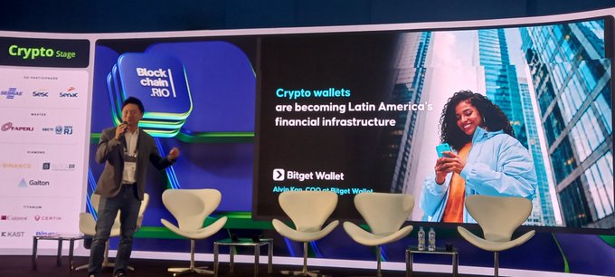 A man gives a presentation at a crypto event with a screen displaying "Blockchain Rio" and a slide titled "Crypto wallets are becoming Latin America's financial infrastructure" featuring Bitget Wallet.