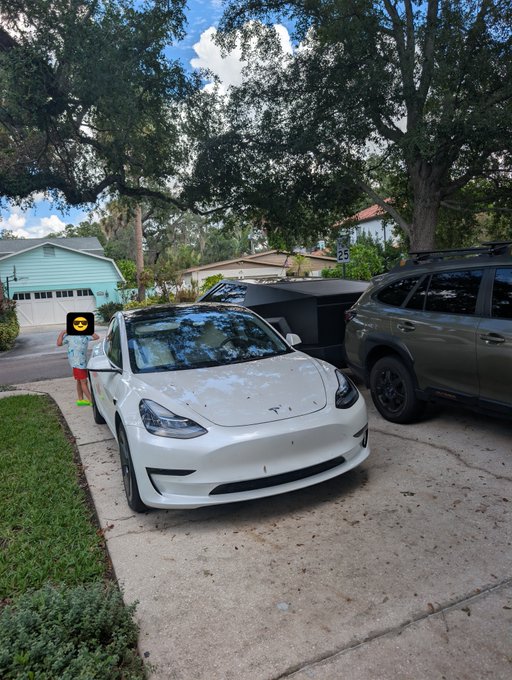 A white Tesla Model 3 parked in a residential driveway with its front trunk open, while a child stands nearby next to a large yellow smiley face emoji overlay covering their face.
