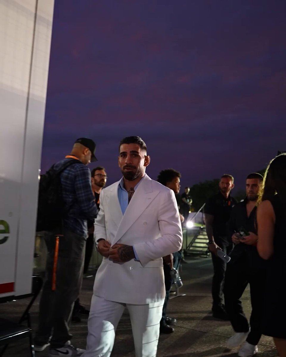 The image shows a man wearing a white suit standing outside, surrounded by several other people who appear to be either waiting or attending an event. The man is dressed in a suit with a tie, giving him a formal appearance. There are at least ten people visible in the scene, some of whom seem to be engaged in conversation while others stand alone. 

The post appears to be related to a UFC fight, as indicated by the use of the hashtag #UFCWhiteHouse Press Conference and the reference to @ParamountPlus. The image also contains text that reads "Only one can leave with the belt @TopuriaIlia vs @Justin_Gaethje have arrived to the !" This suggests that there might be a special event or gathering taking place, possibly related to the UFC fight.