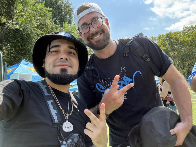Two smiling men pose outdoors under a sunny sky, both making hand gestures while wearing black t-shirts.