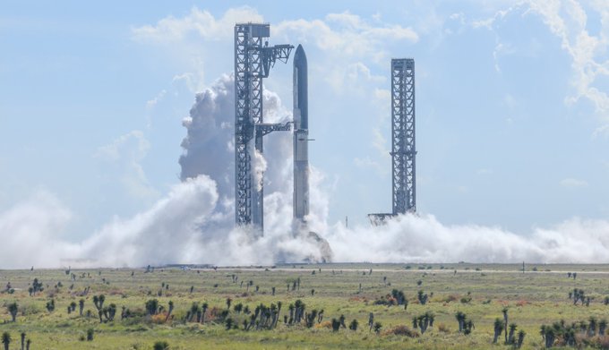 A SpaceX rocket lifts off from a launch pad, with massive plumes of white smoke billowing around the launch tower against a cloudy blue sky.