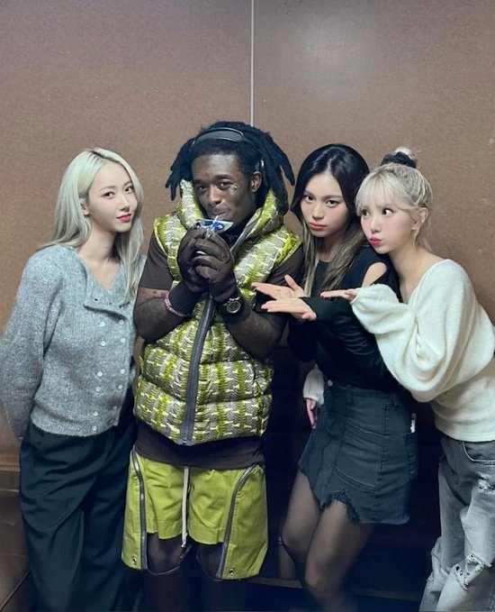 A group photo of five people posing together indoors, including a man in a green patterned puffer vest with dreadlocks in the center, flanked by four women in stylish casual outfits.