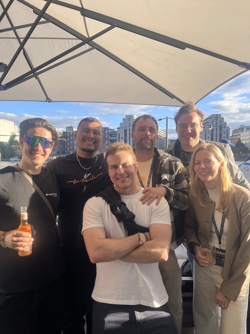 In the image, there are five people standing together under an open umbrella outside of a building. They appear to be friends or colleagues enjoying their time together during a social event. Two men are posing for a group photo with smiles, while two women join in on the fun and stand in front of them. The group seems to be having a good time, creating a lively and memorable atmosphere at the gathering. This type of photograph would be fitting for sharing a moment from an interesting or enjoyable event, such as a meeting like Solana Summit Germany.