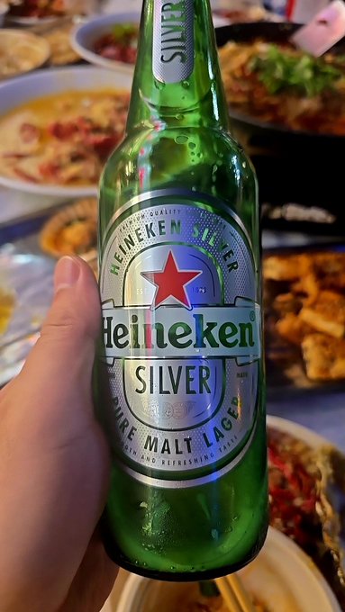 A hand holding up a green bottle of "Heineken SILVER" ("PURE MALT LAGER") in front of a table covered with various plates of food.