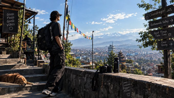 A person with a backpack and cap stands overlooking a sprawling mountain city, with a sleeping dog nearby and directional signs pointing to 'Chandragiri', 'Kathmandu Valley', and other locations.