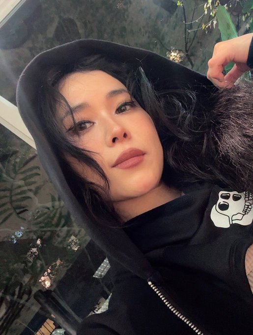 A close-up selfie of an Asian woman wearing a black hooded sweatshirt featuring a white skull graphic, posing against a glass wall with greenery visible behind her.