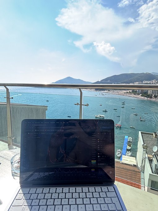A laptop sits on a balcony table overlooking a coastal bay with mountains, boats, and beachgoers visible under a partly cloudy sky.